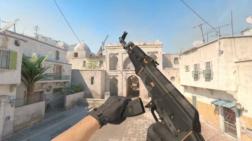 The image presents a first-person view of a player in Counter Strike 2, holding an AK-47 equipped with the "Elite Build" skin. The weapon features a sleek, modern design with black and gold detailing, emphasizing a tactical and refined aesthetic. The background shows a typical Mediterranean-inspired map setting with detailed textures on the buildings and streets, enhancing the realism and immersion of the game environment.