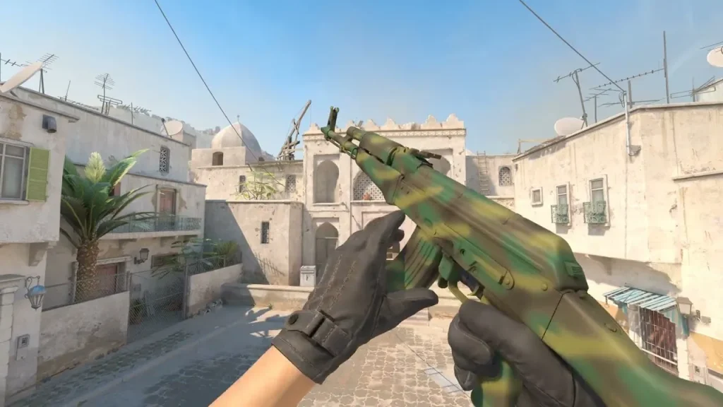 The image illustrates a first-person view of a player in Counter Strike 2 holding an AK-47 with the "Jungle Spray" skin. This skin features a camouflage pattern with shades of green and brown, blending a rough and ready aesthetic suitable for combat in virtual environments resembling outdoor or forested battlefields. The setting shows a sunlit urban map with typical Mediterranean architectural details, providing a stark contrast to the weapon’s jungle-inspired design.