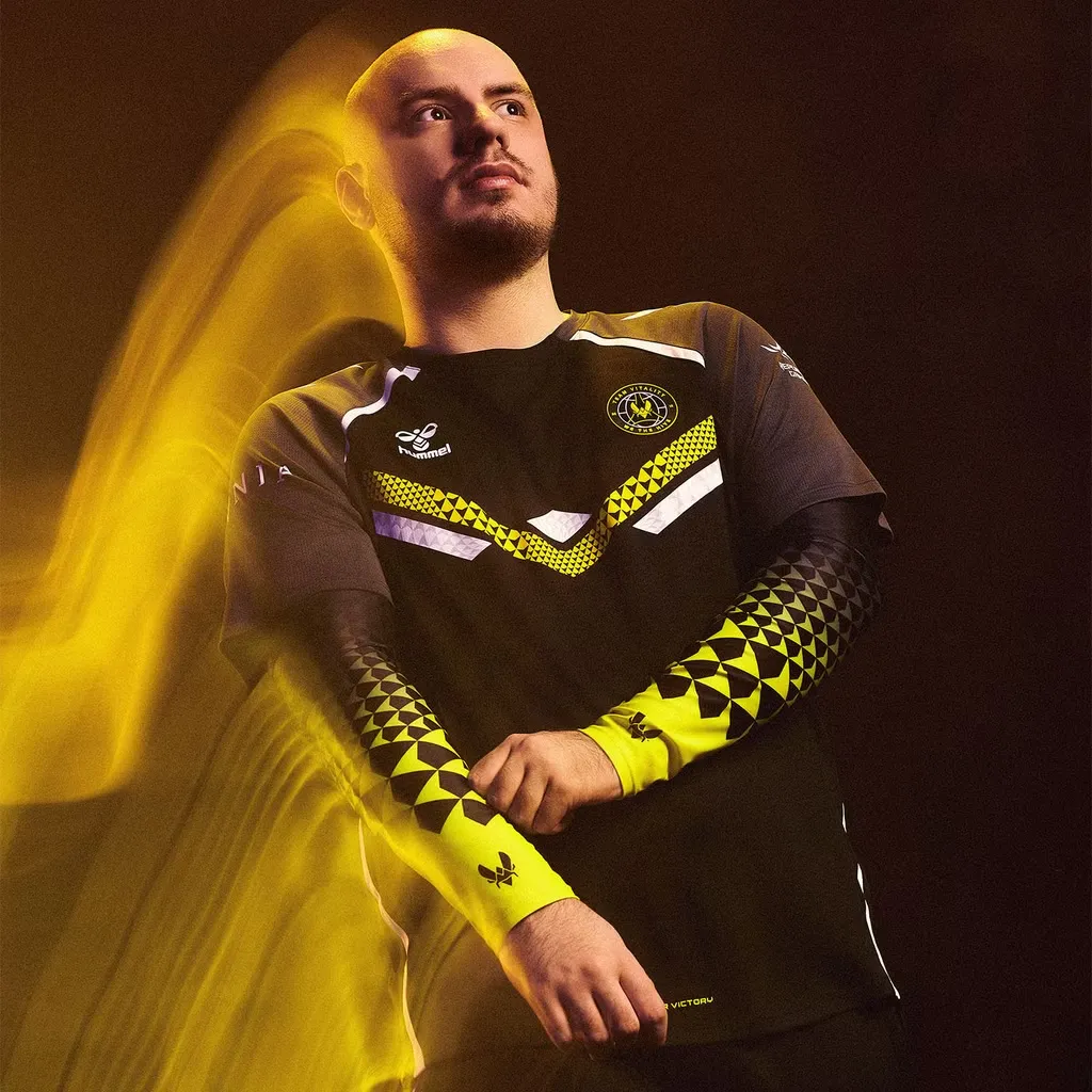 Professional esports player modeling the new Team Vitality pro kit, featuring a stylish black and yellow jersey with geometric patterns. The player is captured in a dramatic pose with a dynamic yellow streak effect, highlighting the modern and energetic design of the kit.