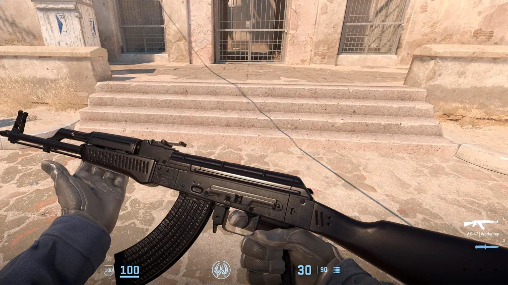 The image showcases a first-person view of the AK-47 | Slate skin in Counter Strike 2, characterized by its minimalist, clean matte black finish. The player is holding the weapon, with a detailed view of the texture and design, which highlights its sleek, professional look. The backdrop is a game environment with a worn stone staircase and a heavy barred door, typical of a competitive map, adding a sense of realism and immersion to the gameplay scene.