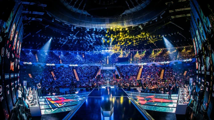 The image captures an electrifying moment at the Intel Extreme Masters (IEM) Katowice, an iconic esports tournament held in a large arena filled to capacity with spectators. The view shows multiple gaming teams competing on stage, surrounded by massive digital displays and vibrant lighting that contribute to a dynamic and immersive viewing experience. The audience, awash in colorful lights, adds to the atmosphere of high-stakes competition, highlighting the event's popularity and significance in the esports world.